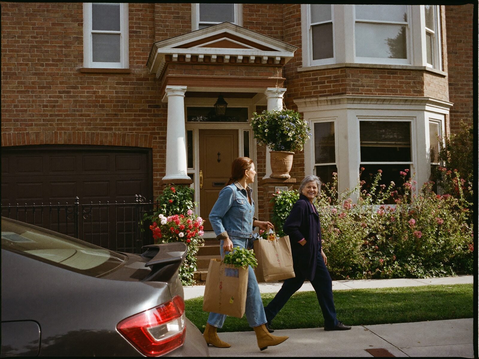 A helper carries grocery bags beside a smiling older woman outside her brick home
