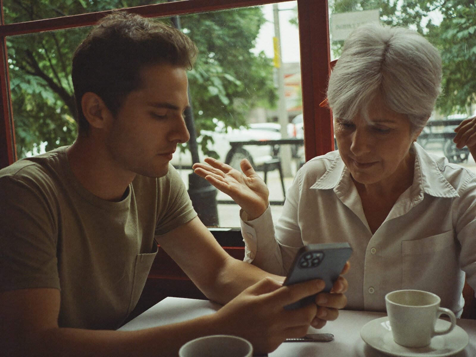 A patient helper and an older woman work through her phone together at a sunny cafe table