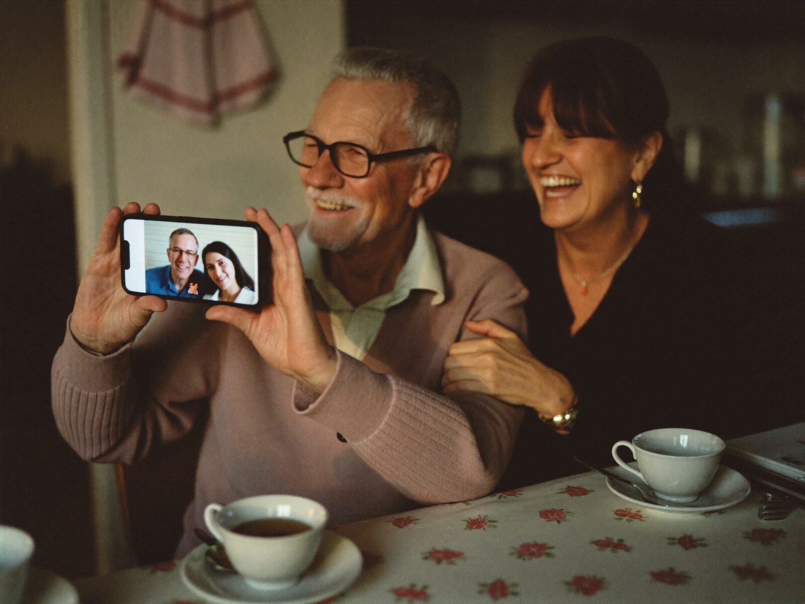 An older man beams at a smartphone video call with his grandchildren while a helper shares the moment