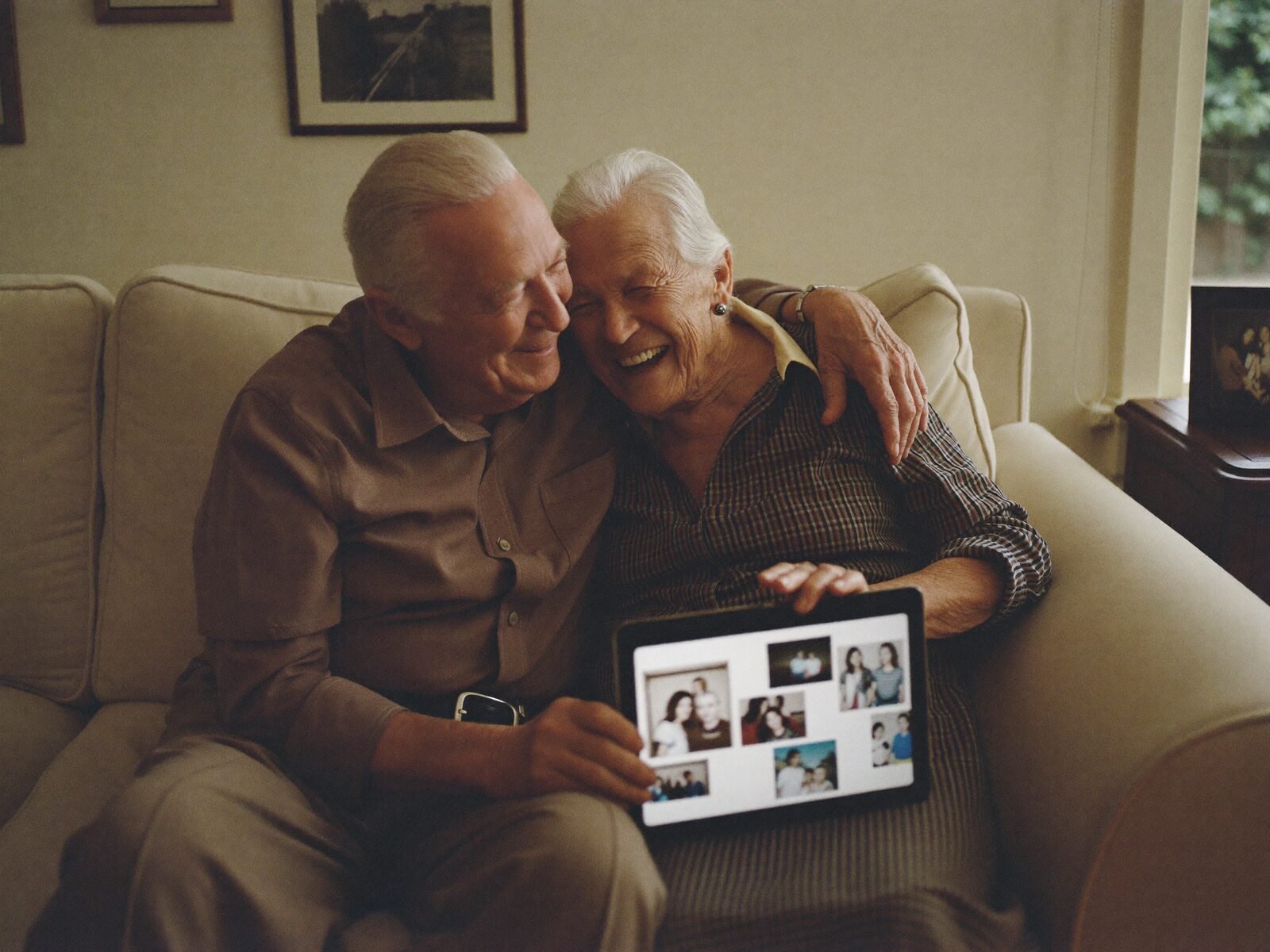 An older couple laugh together on their sofa while looking at family photos on a tablet