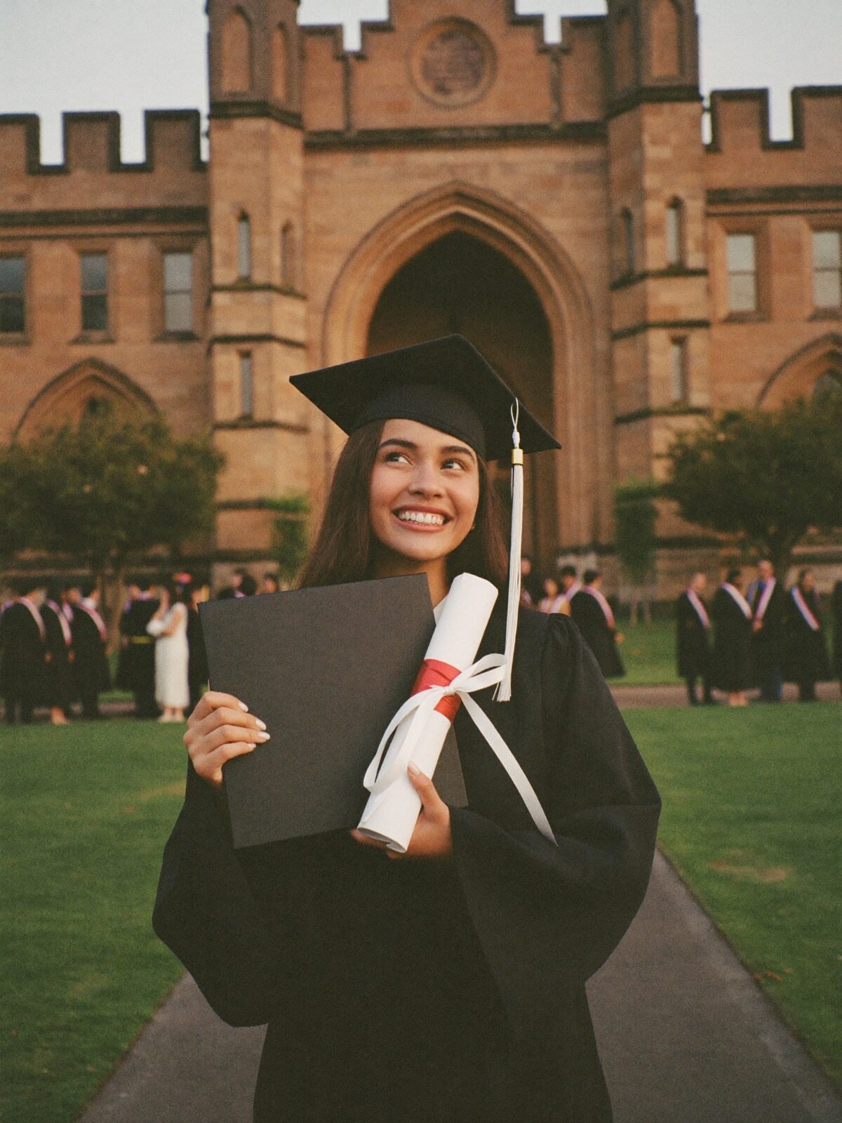A graduate in cap and gown smiles in front of a sandstone university building