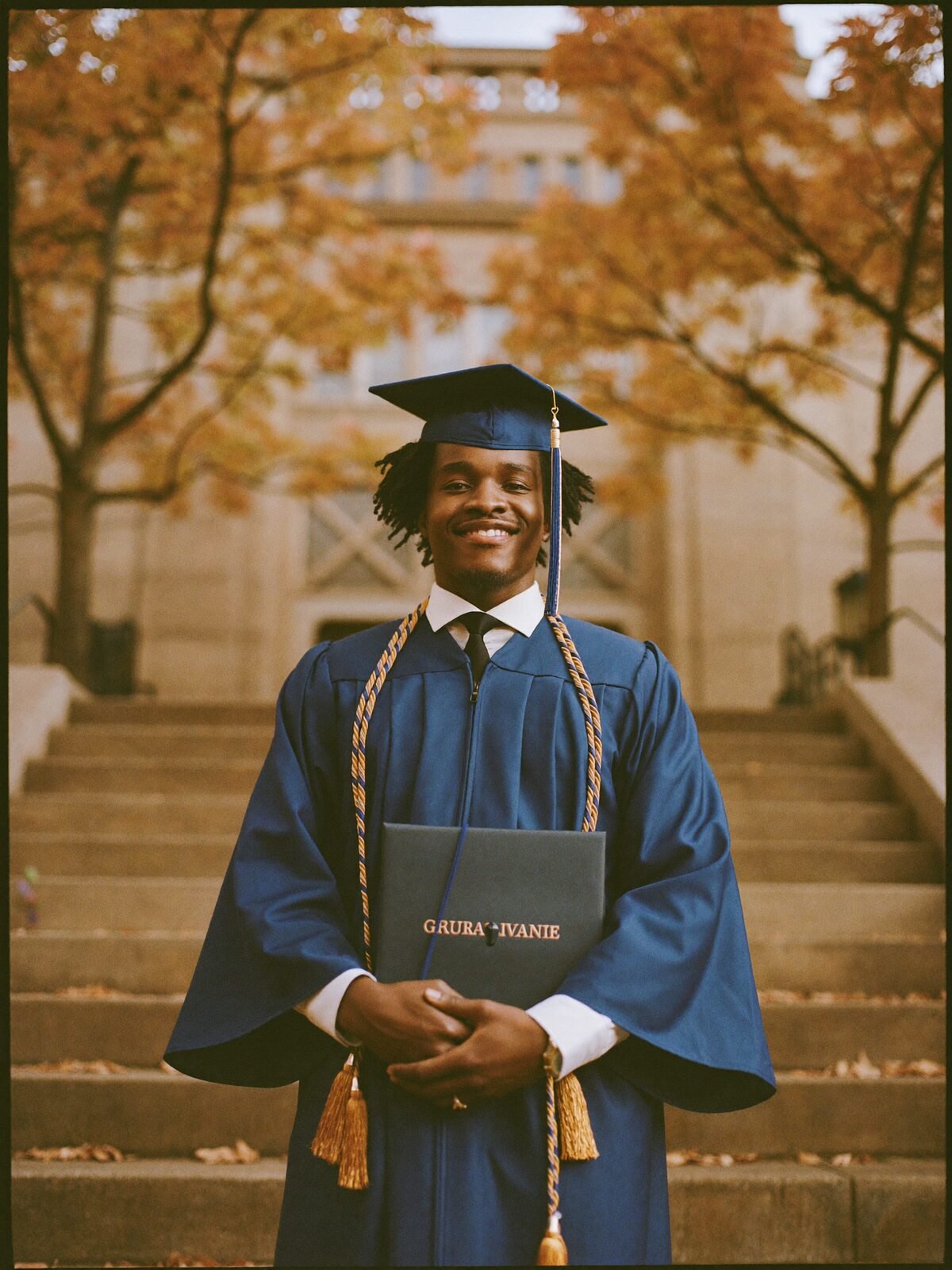 A graduate in a blue gown with a gold stole stands proudly on sandstone university steps