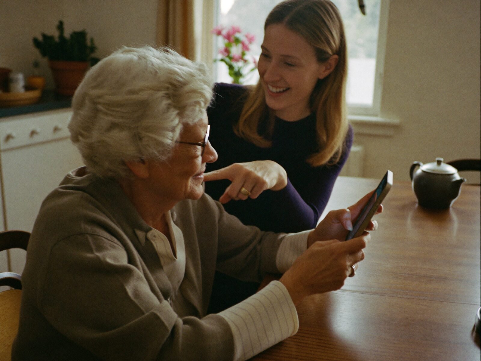 A helper sits beside an older woman at her kitchen table, showing her something on her smartphone