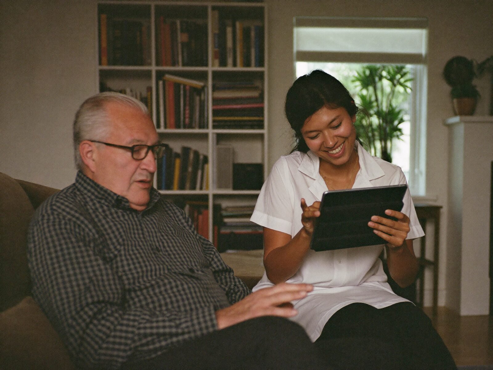 An older man and a young helper smile at a tablet together on a sofa