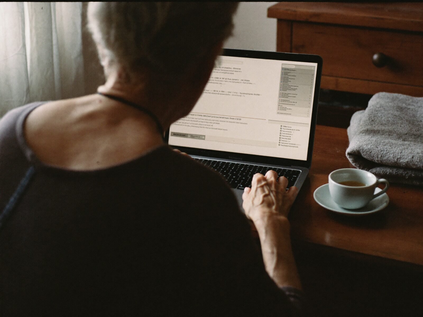 An older woman's hands typing on a laptop at a wooden desk with a cup of tea beside it
